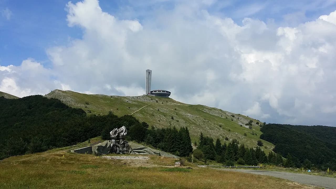 The saucer-shaped Buzludzha monument and its star tower on a green ridge, with the torch monument below
